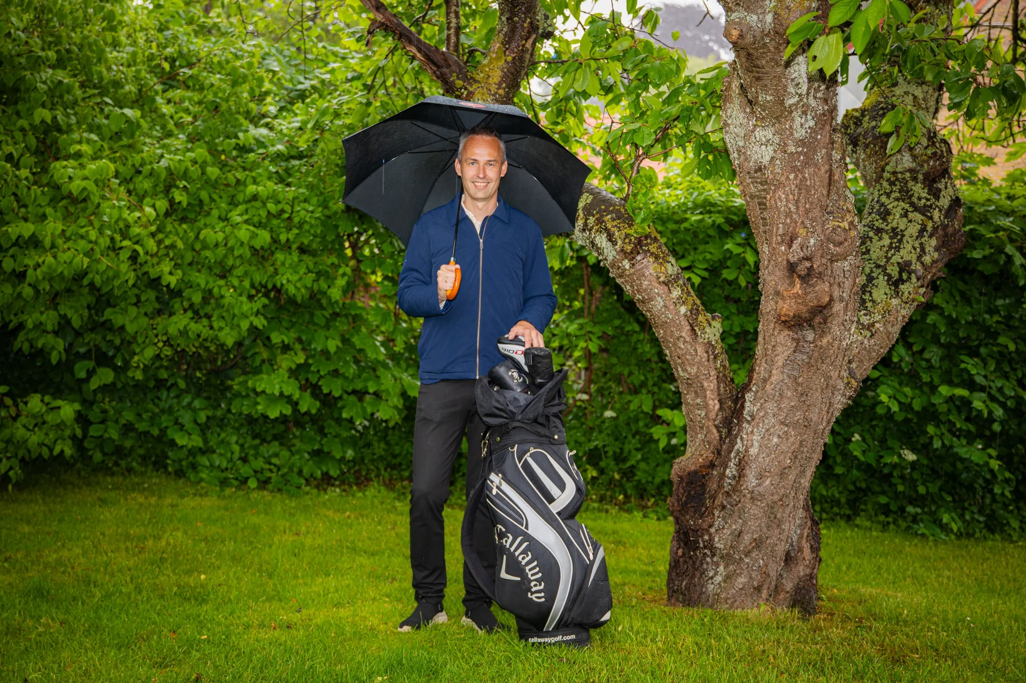 Daniel with an umbrella and his golf bag under a tree